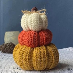 May include: A three-tiered stack of crocheted pumpkins in autumnal hues. The base pumpkin is yellow, the middle one orange, and the top one white. A twine bow and brown stem adorn the white pumpkin. A seasonal decorative item.