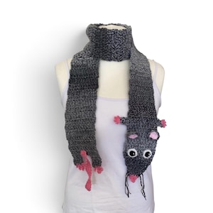 May include: A crocheted scarf featuring a gradient grey yarn texture. One end of the scarf is shaped like a rat head with pink ears, black and white eyes, and black whiskers. The other end features a pink tail and four small pink paws.