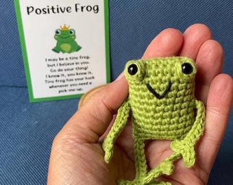 May include: A handmade green crochet frog with black eyes and a smiling mouth. A card with the text "Positive Frog" and a cartoon frog illustration is in the background. The card includes inspirational text.