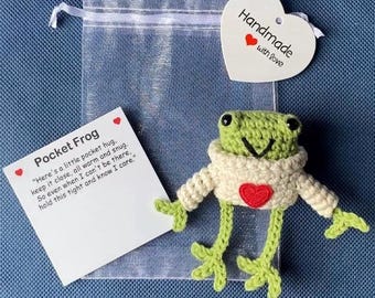 May include: A handmade crochet frog wearing a cream-colored sweater with a red heart. The frog is green with black eyes and a smiling mouth. A tag reads "Handmade with Love." A card says "Pocket Frog" with a poem.
