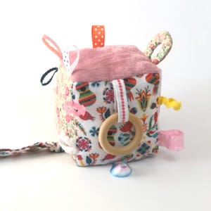 Può includere: Un cubo di tessuto rosa e bianco con motivi e trame colorate. Ha un anello di legno, nastri e un campanello attaccato. Questo è un giocattolo sensoriale per bambini.