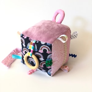 Può includere: Cubo di attività per bambini rosa e nero con un motivo arcobaleno, un anello di legno e vari nastri e trame.