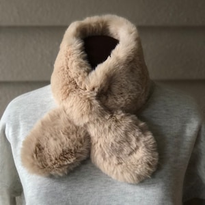 以下が含まれることがあります： ベージュのフェイクファーのストールで、幅広でふわふわの襟が付いています。ストールは半分に折り畳まれ、マネキンの首にかけられています。
