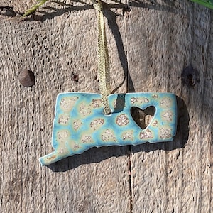 May include: A blue and white ceramic ornament shaped like the state of Connecticut with a heart cut out in the center. The ornament is hanging from a gold ribbon.