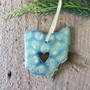 May include: A blue and green ceramic ornament shaped like the state of Ohio with a heart cut out in the center. The ornament is hanging from a gold ribbon.