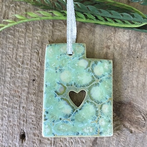 May include: A green ceramic ornament shaped like the state of Utah with a cutout heart in the center. The ornament has a white string for hanging.