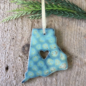May include: A blue and green ceramic ornament shaped like the state of Alabama with a heart cut out in the center. The ornament has a gold ribbon attached for hanging.