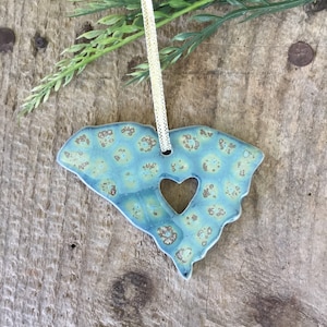 May include: A blue and green ceramic ornament shaped like the state of South Carolina with a heart cut out in the center. The ornament has a gold string attached to it.