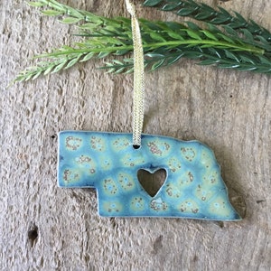 May include: A blue and brown ceramic ornament shaped like the state of Nebraska with a heart cut out in the center. The ornament is hanging from a gold ribbon.