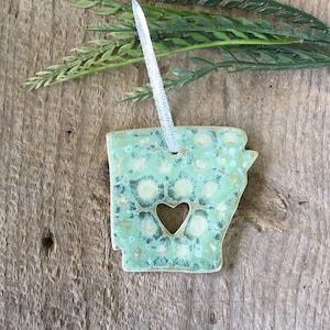 May include: A green and white ceramic ornament shaped like the state of Arkansas with a heart cut out in the center. The ornament has a silver string for hanging.