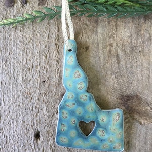 May include: A blue and green ceramic ornament shaped like the state of Idaho with a heart cut out in the center. The ornament has a white string for hanging.