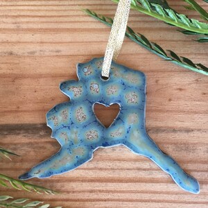 May include: A ceramic ornament in the shape of Alaska, with a heart-shaped cutout. The ornament has a blue and tan mottled glaze and a gold ribbon for hanging. Decorative item.