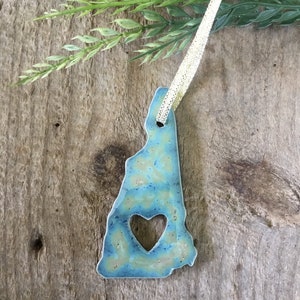 May include: A blue ceramic ornament shaped like the state of New Hampshire with a heart cut out in the center. The ornament is hanging from a gold ribbon.