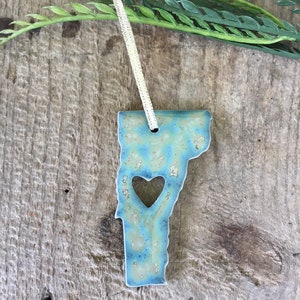 May include: A blue and green ceramic ornament shaped like the state of Vermont with a heart cut out in the center. The ornament is hanging from a gold string.