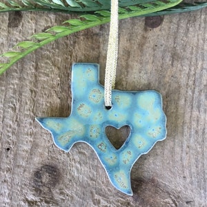 May include: A blue and green ceramic ornament shaped like the state of Texas with a heart cut out in the center. The ornament has a gold string attached to it.