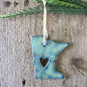 May include: A blue and green ceramic ornament shaped like the state of Minnesota with a heart cut out in the center. The ornament is hanging from a gold ribbon.
