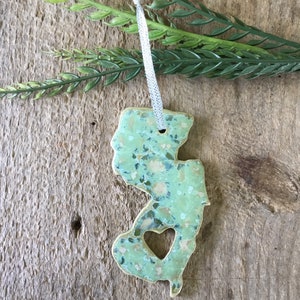 May include: A green and white ceramic ornament shaped like the state of New Jersey with a heart cut out in the center. The ornament has a white ribbon attached for hanging.