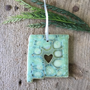 May include: A green ceramic ornament shaped like the state of New Mexico with a heart cut out in the center. The ornament has a white string for hanging.