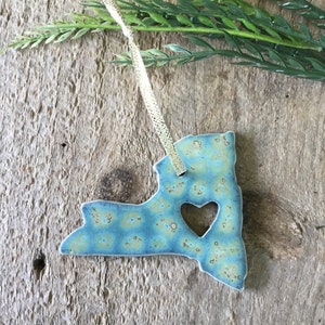 May include: A blue and green ceramic ornament shaped like the state of New York with a heart cut out in the center. The ornament is hanging from a silver string.