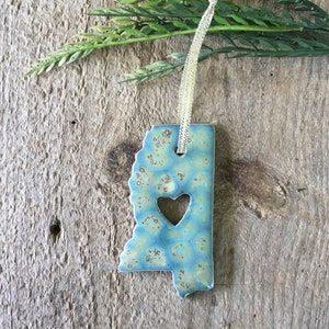 May include: A blue and green ceramic ornament shaped like the state of Mississippi with a heart cut out in the center. The ornament is hanging from a white ribbon.