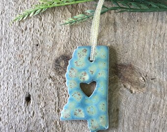 May include: A blue and green ceramic ornament shaped like the state of Mississippi with a heart cut out in the center. The ornament is hanging from a white ribbon.