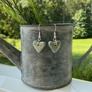May include: A pair of heart-shaped earrings with a teal glaze and brown floral accents. The earrings hang from silver-colored hooks. The earrings are displayed in a galvanized metal watering can.