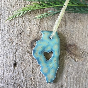 May include: A blue and yellow ceramic ornament shaped like the state of Illinois with a heart cut out in the center. The ornament is hanging from a gold ribbon.