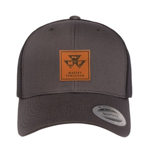 May include: A dark gray trucker hat with a brown leather patch featuring the Massey Ferguson logo. The logo is a stylized tractor with the letters "MF" in the center. The hat has a black mesh back.