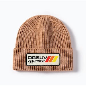 May include: A brown ribbed knit beanie with a rectangular patch. The patch features the text "OGSUV RUNNER" in white and black, with red, orange, and yellow stripes. The beanie is a warm, casual accessory.
