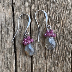 May include: A pair of dangle earrings featuring faceted, teardrop-shaped gray labradorite stones topped with clusters of small, round, deep pink ruby beads. The stones are secured with silver-toned wire and hang from curved silver-toned ear wires.