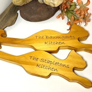 May include: Two wooden cat-shaped kitchen utensil holders. The top holder is engraved with "The Baumgarts Kitchen" and the bottom holder is engraved with "The Stapletons Kitchen".