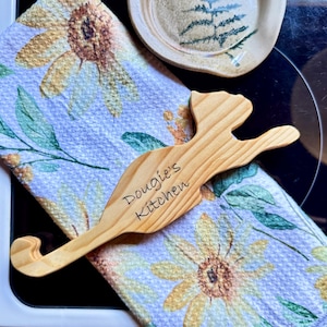 May include: A light-colored wooden spoon rest shaped like a cat with the text "Dougie's Kitchen" engraved on the surface. It is positioned on a folded kitchen towel featuring a yellow and white sunflower pattern with green leaves.