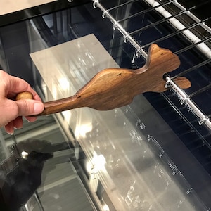May include: A wooden spatula with a unique, curved design. The spatula is brown and has a natural wood finish. It is resting on a metal rack inside an oven.