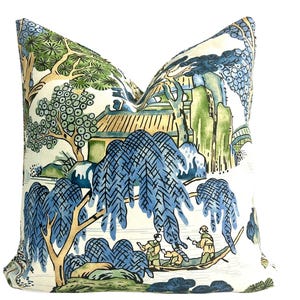 May include: Decorative throw pillow with a scenic landscape design. The pillow features a white background with a detailed illustration of a blue and green Asian-inspired landscape, including trees, a bridge, and figures in a boat.