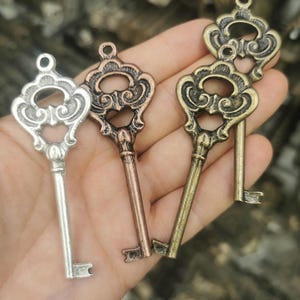 Puede incluir: Tres dijes de llave de metal con cabezas ornamentadas de volutas y vástagos simples. Un dije es de tono plateado, otro de tono cobre y otro de tono latón. Cada llave tiene un pequeño lazo circular en la parte superior para sujetar.