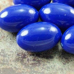 May include: A group of six smooth, oval-shaped beads in a vibrant blue color. The beads are arranged in a cluster on a textured, brown surface.