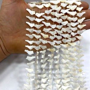 May include: Strands of white bird-shaped beads. The beads are strung together on clear string, creating a delicate and airy appearance. The beads are held in a hand, showcasing their size and detail. Ideal for jewelry making or craft projects.