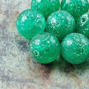 May include: A group of green glass marbles with a sparkly finish. The marbles are scattered on a textured surface.