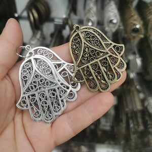 Puede incluir: Dos dijes de mano de Hamsa con intrincados recortes de filigrana. Un dije tiene un acabado en tono plateado brillante, mientras que el otro tiene un acabado en tono bronce antiguo. Cada dije incluye una pequeña argolla de metal en la parte superior.