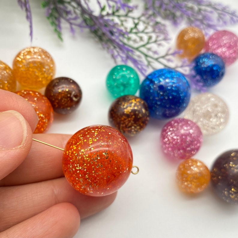 Puede incluir: Surtido de cuentas redondas de colores con purpurina incrustada en resina transparente. Las cuentas son de varios colores, incluyendo naranja, marr&oacute;n, azul, verde, rosa y transparente.