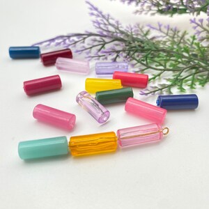 May include: Assortment of colorful glass beads in various shades of pink, blue, yellow, and green. The beads are cylindrical in shape and are arranged on a white surface.