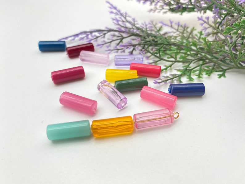 May include: Assortment of colorful glass beads in various shades of pink, blue, yellow, and green. The beads are cylindrical in shape and are arranged on a white surface.