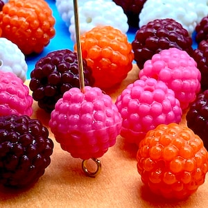 May include: A selection of colourful, round beads with a textured surface. The beads are in shades of orange, pink, white, and dark red. A gold-coloured wire is inserted into one of the pink beads. The background is a mix of blue and tan.