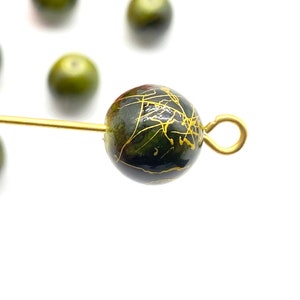 May include: A dark green glass bead with gold accents and a gold loop attached. The bead has a marbled effect.