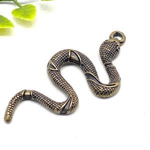 May include: A bronze-colored snake charm with a textured, scaled design. The snake is curled in a sinuous shape, with a loop at the head for attaching to jewelry or other crafts. The charm is set against a white background.