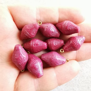 May include: A collection of ten maroon-colored, barrel-shaped beads with intricate embossed patterns. Each bead has a small gold-colored loop at the top for stringing. The beads are displayed against a light background.