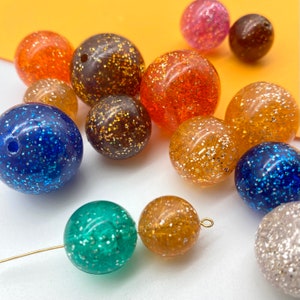 Puede incluir: Surtido de cuentas redondas llenas de purpurina de varios colores, incluyendo azul, verde, naranja, marr&oacute;n y plateado.