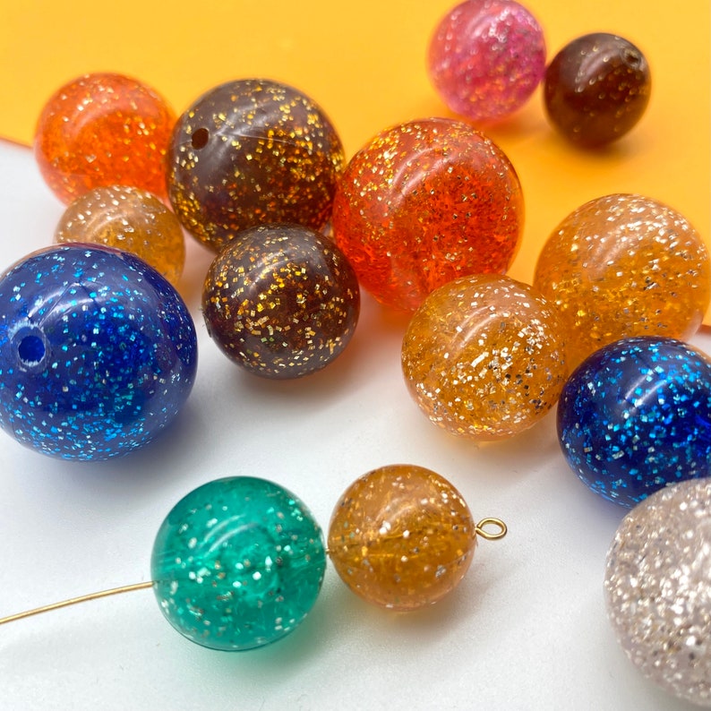 Puede incluir: Surtido de cuentas redondas llenas de purpurina de varios colores, incluyendo azul, verde, naranja, marr&oacute;n y plateado.