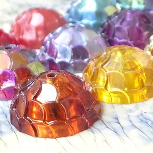 Könnte beinhalten: Sortiment von bunten, irisierenden, kuppelförmigen Cabochons mit einer strukturierten, schuppigen Oberfläche. Die Cabochons sind aus Kunststoff und haben einen Durchmesser von etwa 2,5 cm.