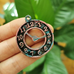 Puede incluir: Un dije de reloj redondo y desgastado con esfera negra, borde rosa y detalles turquesa. Los números del 1 al 12 son visibles. Las manecillas del reloj son turquesas. El dije tiene un pequeño lazo en la parte superior.